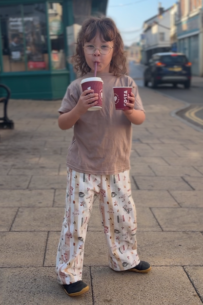 A young girl stands on a paved street holding two red cups, wearing a beige t-shirt and patterned pajama pants. Buildings and a shop sign are visible in the background.